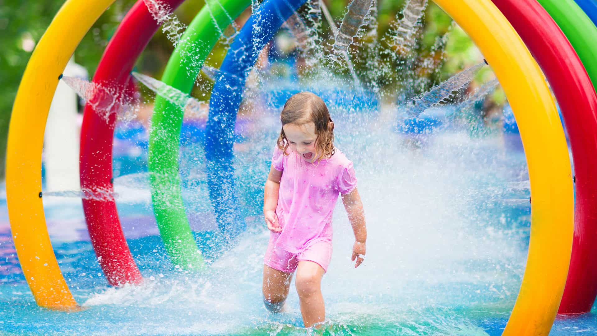 Happy toddler girl running through a water fountain in a swimming pool enjoying water amusement park while on summer vacation
