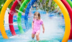 Happy toddler girl running through a water fountain in a swimming pool enjoying water amusement park while on summer vacation