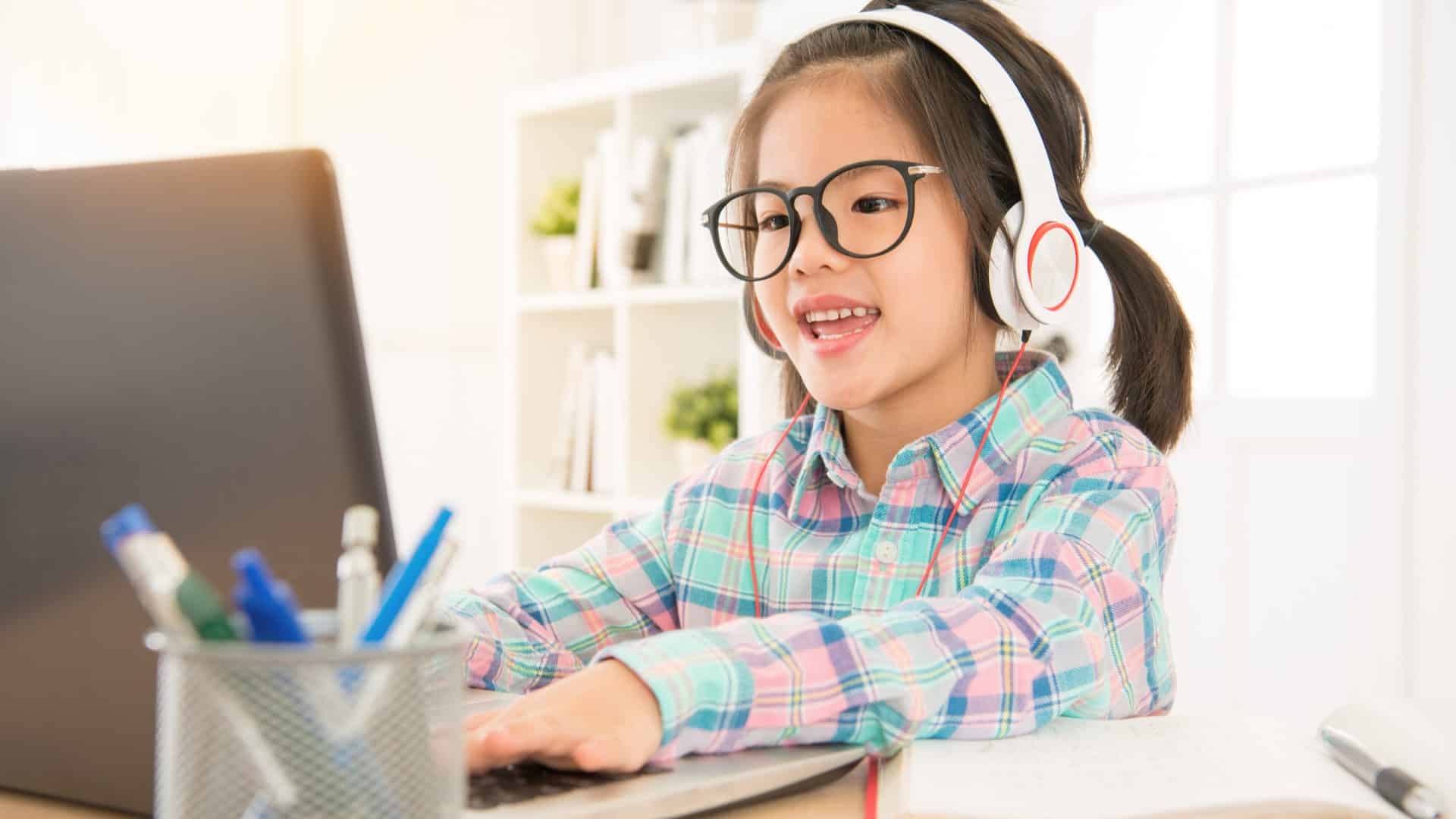 Girl with Asian roots sitting in front of a computer doing elearning and online classes using headsets